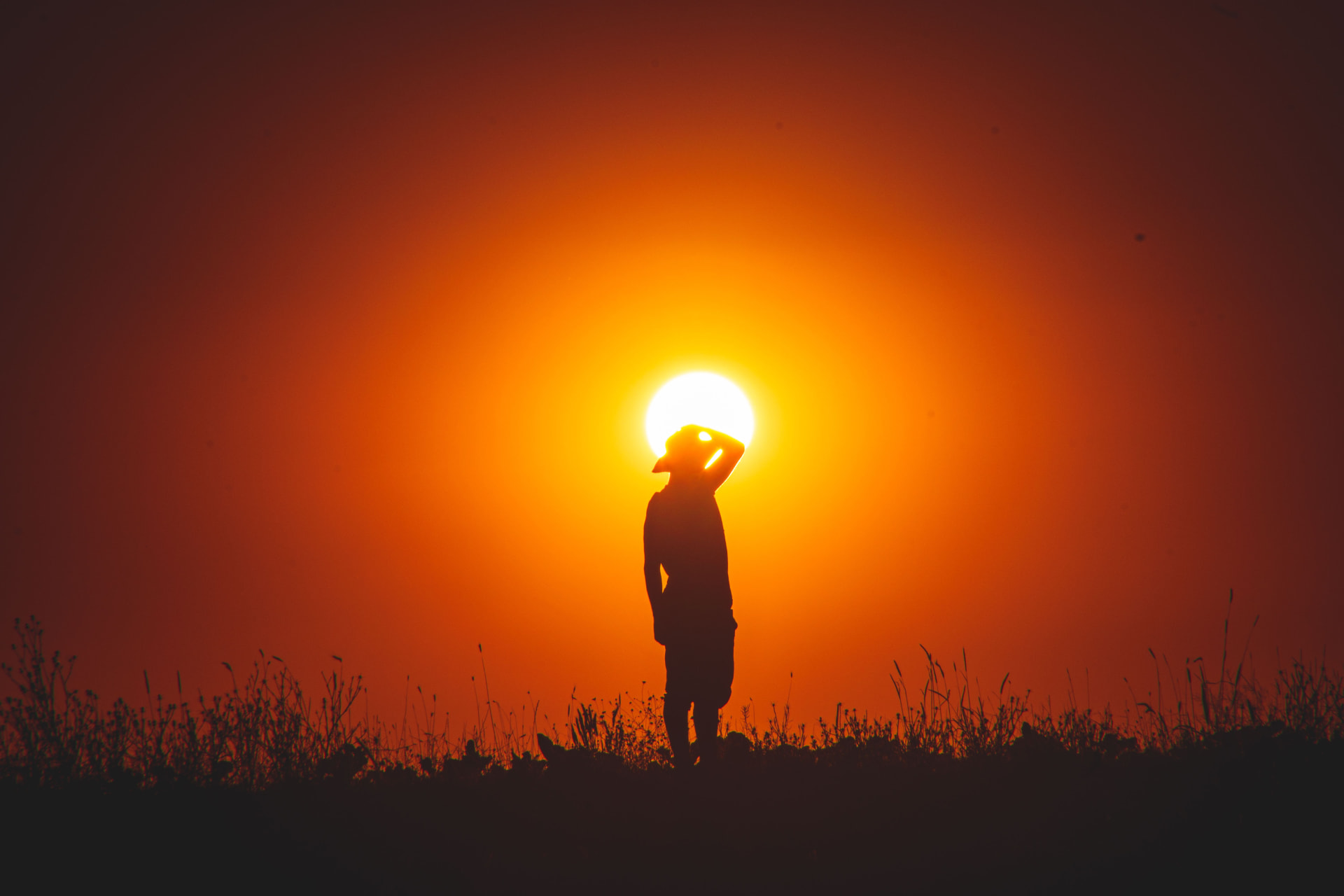 Silhouette of a person standing in a field against a blazing orange sunset, shielding their eyes from the sun